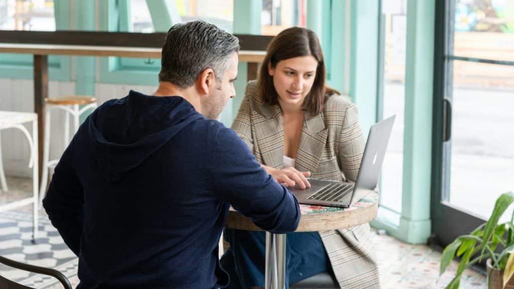 A woman in a plaid blazer uses a laptop while talking to a seated man.