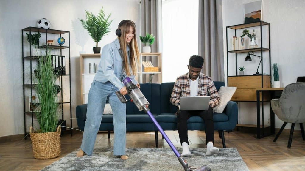 A woman wearing headphones is cleaning the carpet while her husband is working on a laptop.