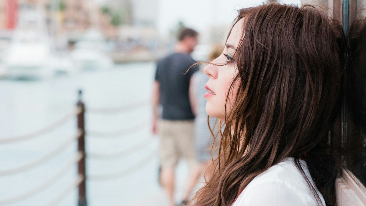 A woman with long brown hair looks away while leaning against a wall outdoors near water.