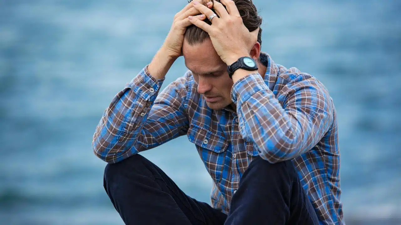 A man in a plaid shirt sits with his head down and hands on his forehead.