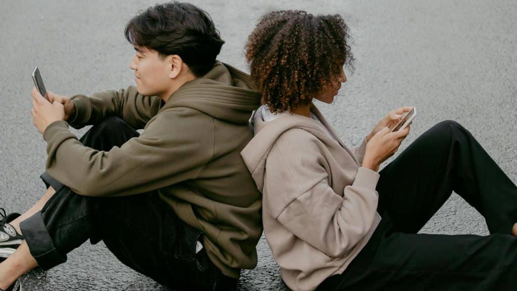 A pair of people sitting back-to-back on the ground, each focused on their phone.