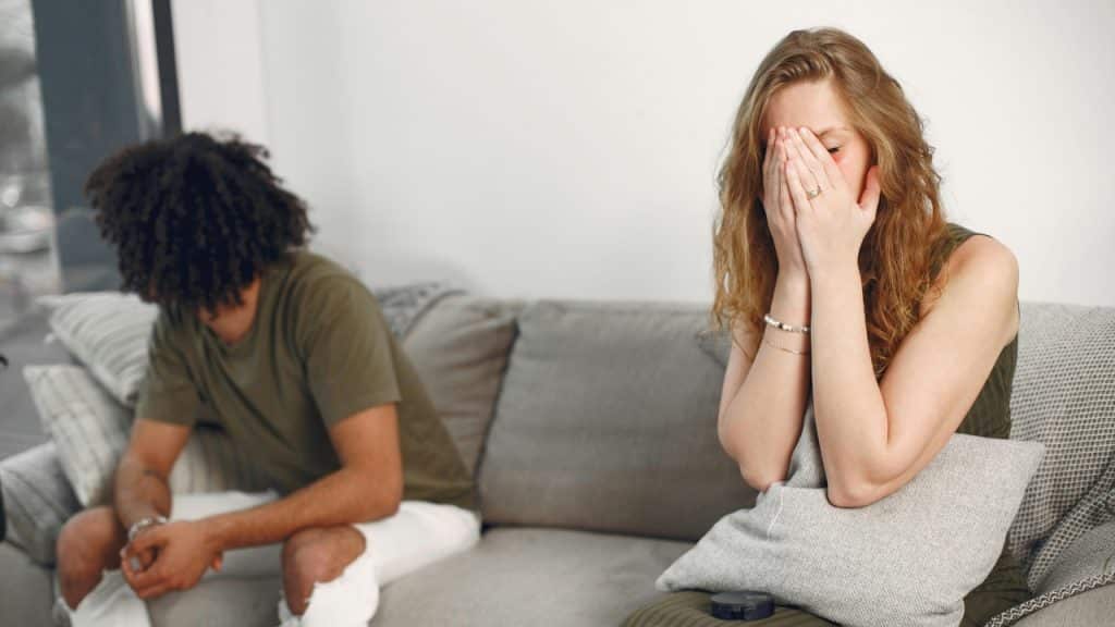A woman sits on a couch covering her face while a man sits looking away.