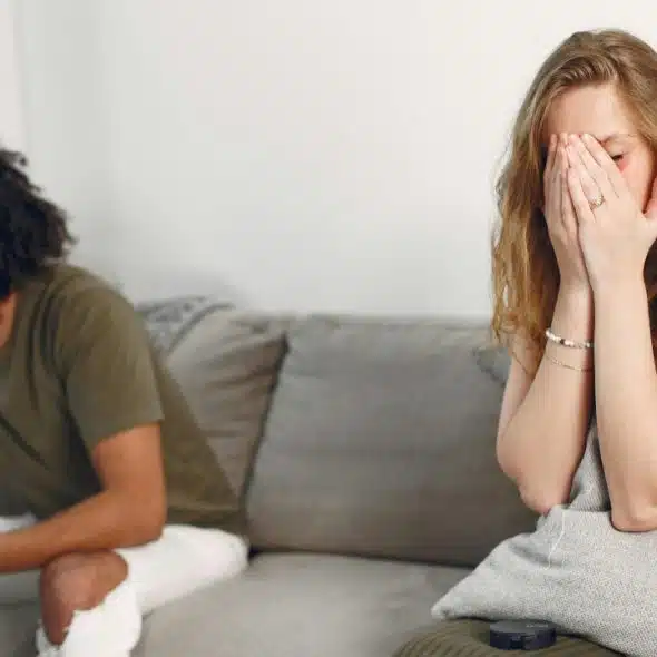 A woman sits on a couch covering her face while a man sits looking away.