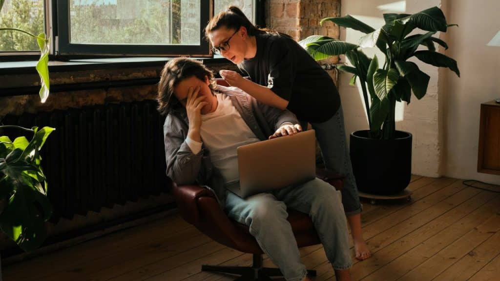 A woman stands over a man sitting in a chair with his hand on his forehead.