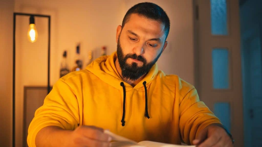 A bearded man in a yellow hoodie sits at a table reading a book under warm lighting.