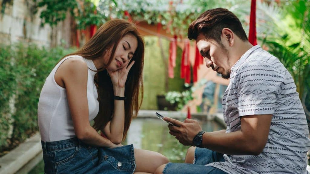 A woman rests her head on her hand while a man beside her looks at a phone.