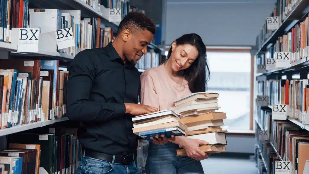 A man and woman stand between library bookshelves smiling while looking at a large stack of books.