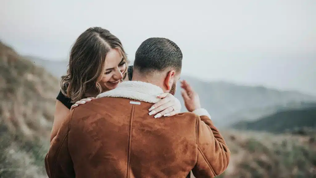 A couple embracing outdoors, with a woman smiling over a man’s shoulder.