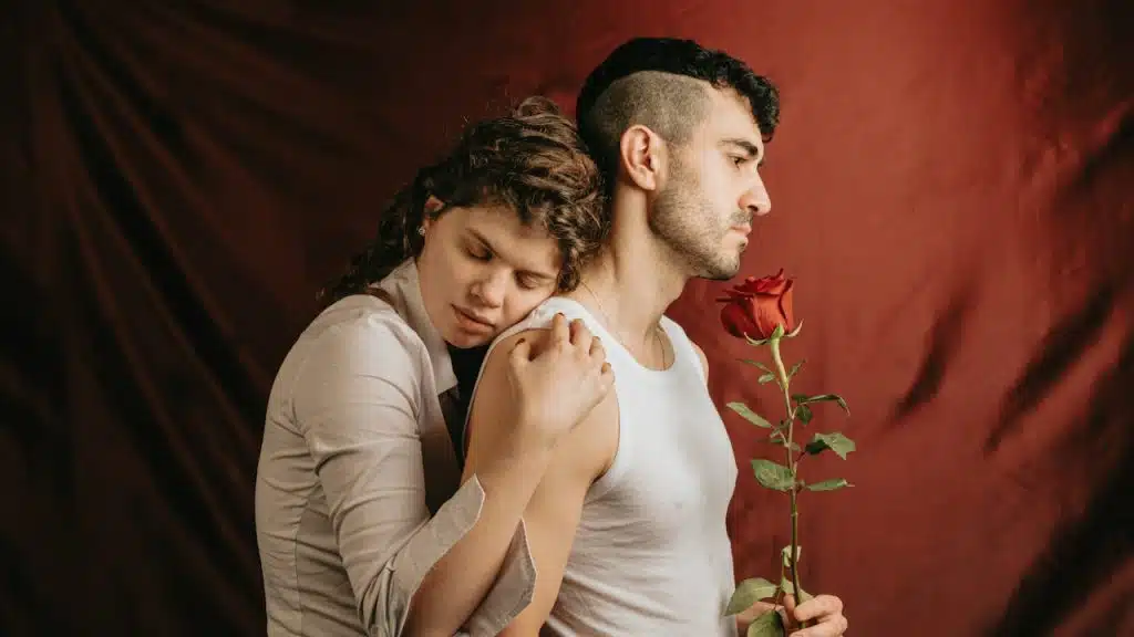 A man is holding a rose, and a woman is hugging him from behind.