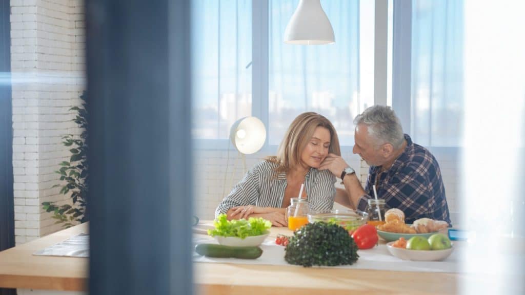 An older man affectionately touches a woman's face as they sit together at a dining table with fresh food in a sunlit room.
