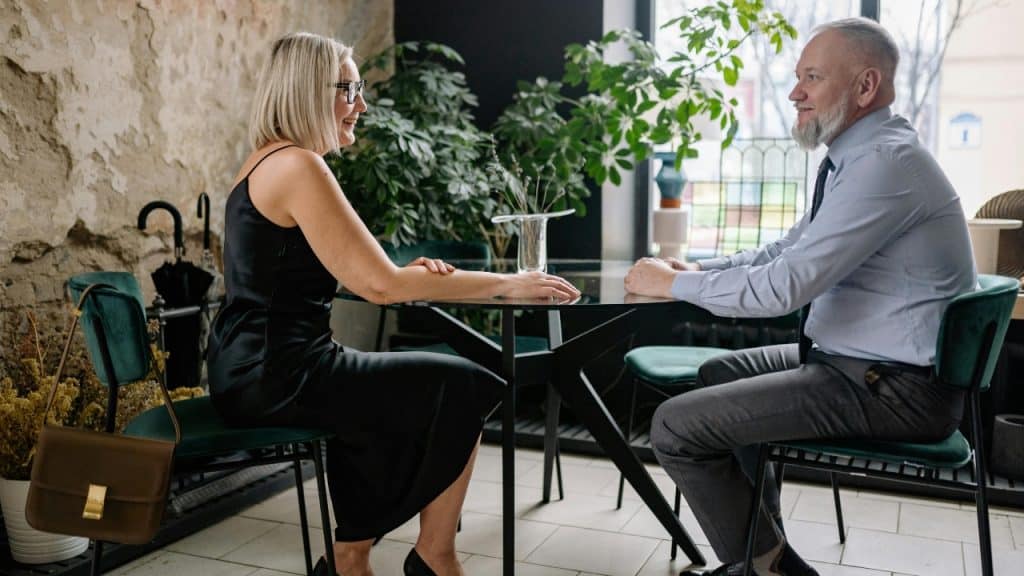 A blonde woman and an older man sit at a glass table in a cafe setting.