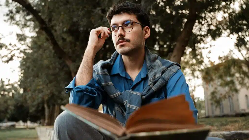 A man with a mustache and glasses sits outdoors holding an open book and looking up.