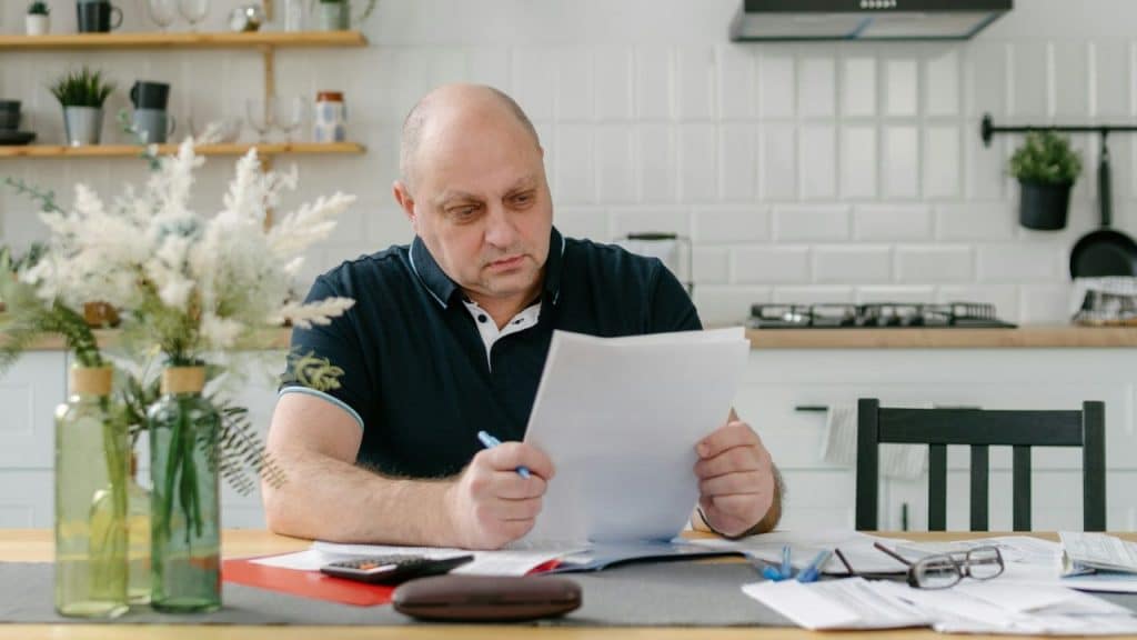 A man sits at a kitchen table reviewing several white papers and financial documents.