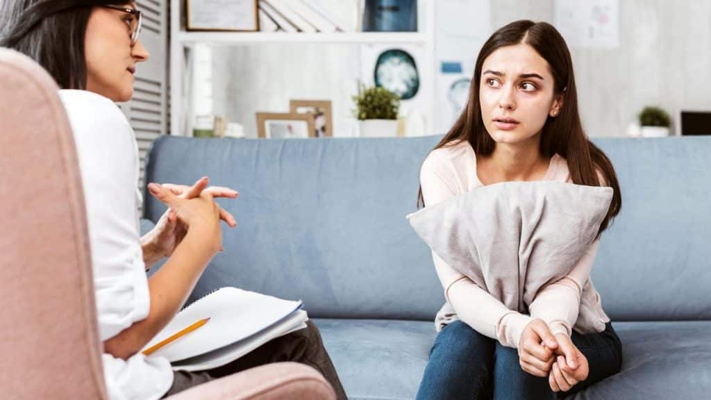 A worried woman clutching a pillow while talking to another person in a counseling session.