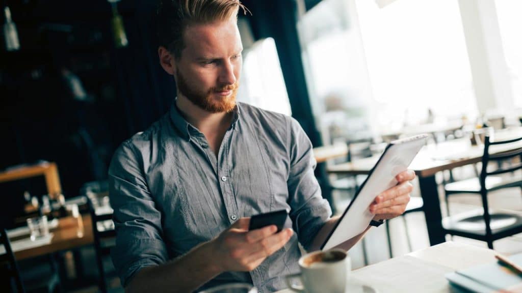 A man sitting in a café, looking at a tablet while holding a phone.