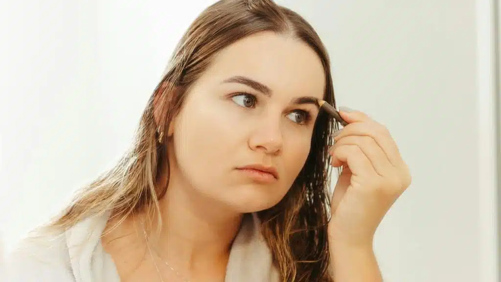 A woman grooming her eyebrow with a pencil while looking off to the side.