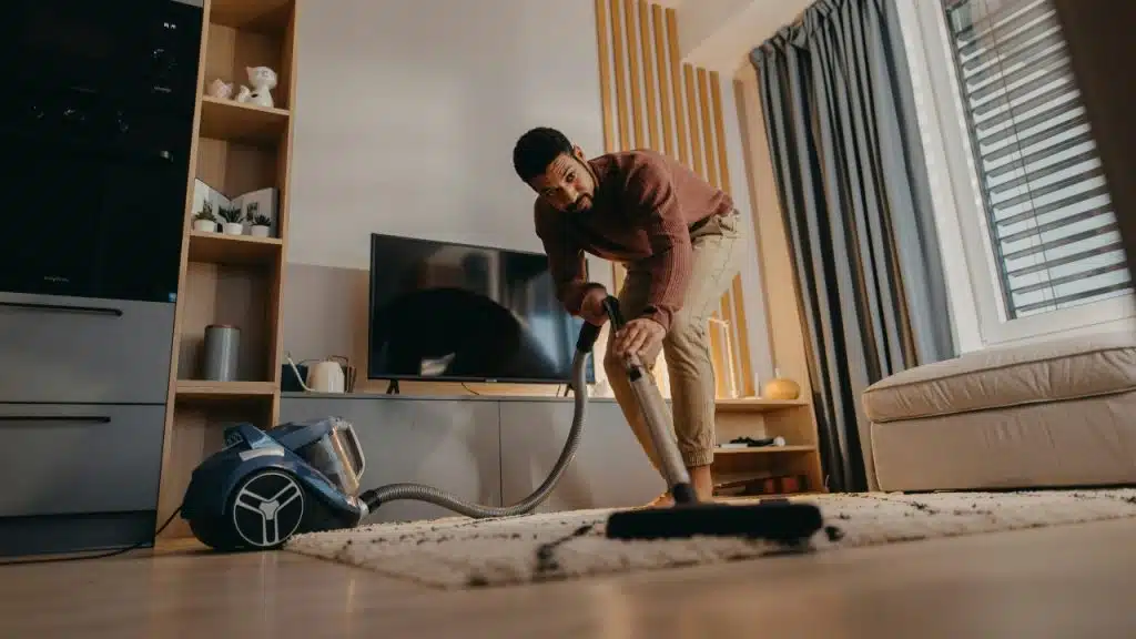 A man in a brown sweater uses a vacuum cleaner on a rug indoors.