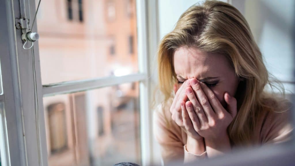 A blonde woman covers her face with both hands while sitting near a window.