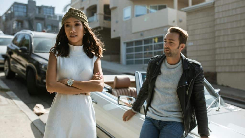 A woman in a white dress stands with crossed arms near a man and car.