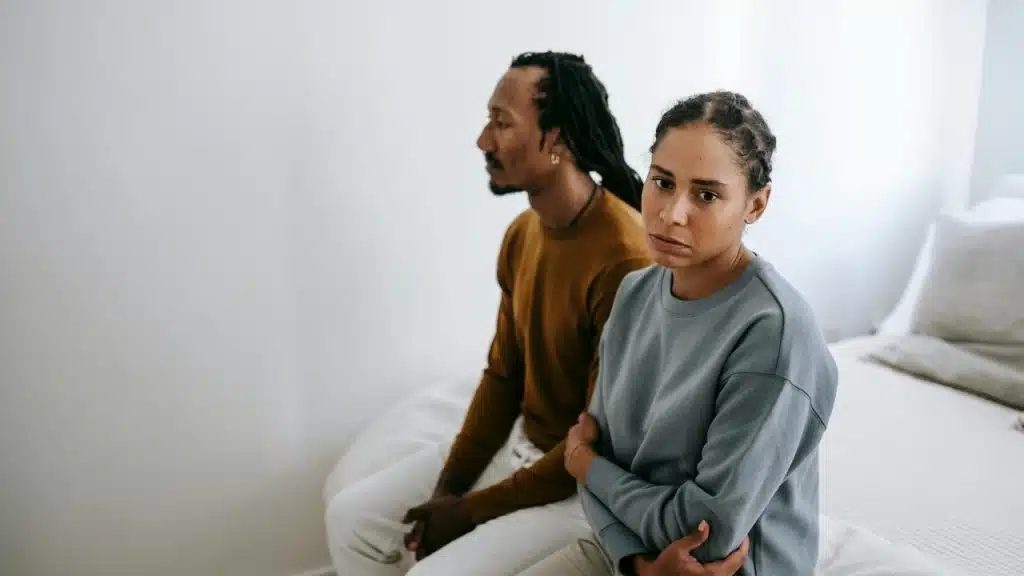 A man and woman sitting on a bed looking in different directions with neutral expressions.
