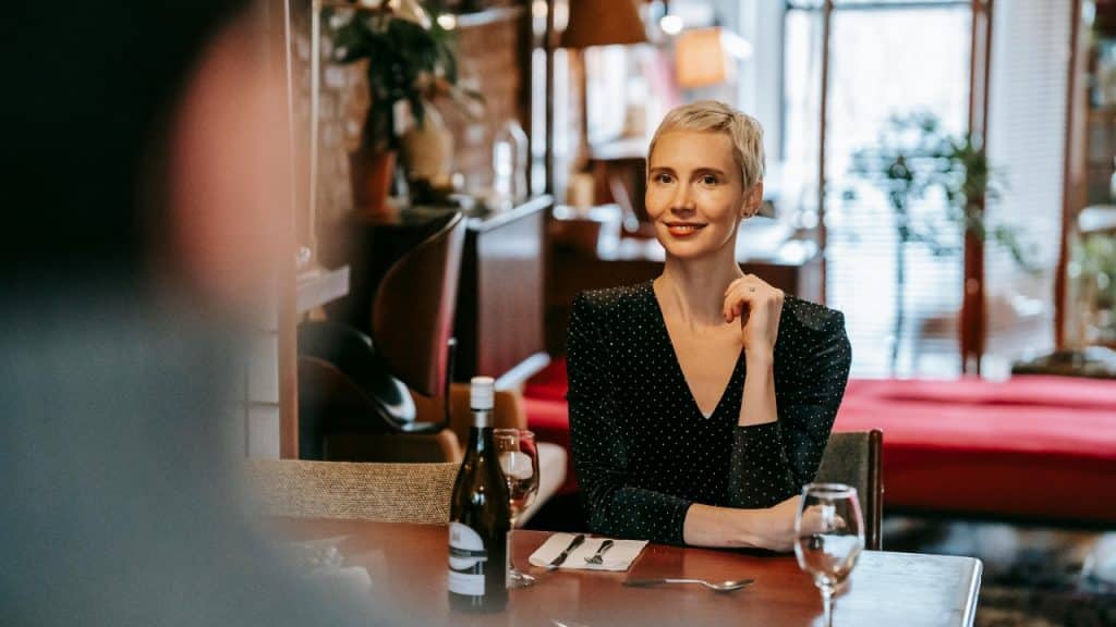 A woman with short blonde hair sits at a restaurant table looking toward another person.