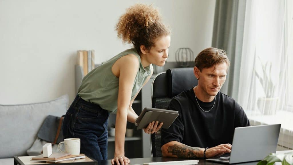 A woman leans over a desk with a tablet while a man sits and types on a laptop.