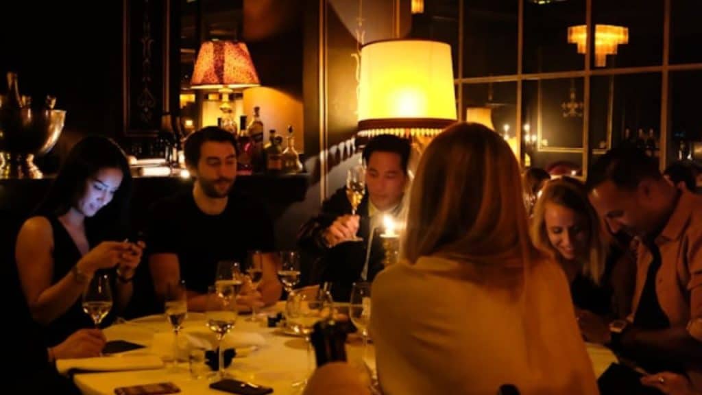 Several people sit around a circular dinner table in a dimly lit restaurant with warm lamps.