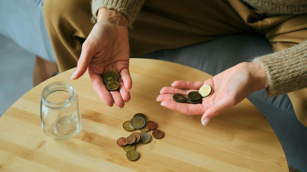hands counting coins near a glass jar on a table.