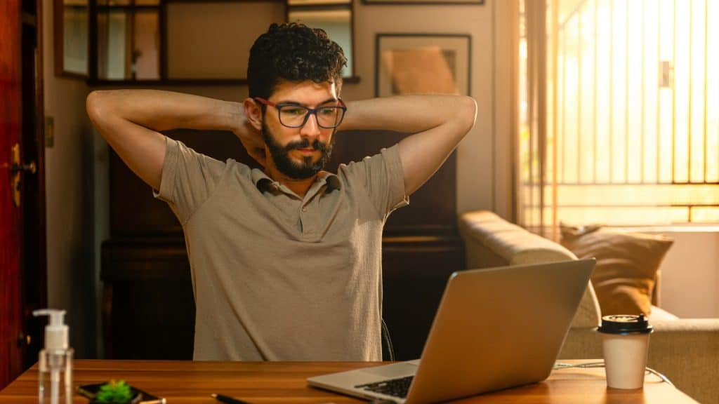 A man with glasses and curly hair sits at a desk with his hands behind his head.