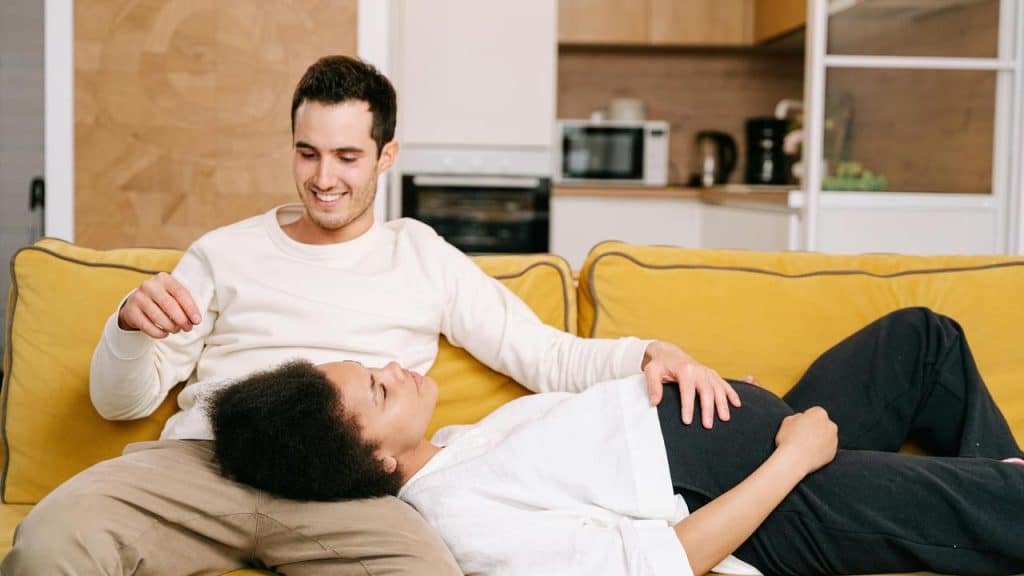 A couple relaxing on a couch, with a pregnant woman lying across a man’s lap.
