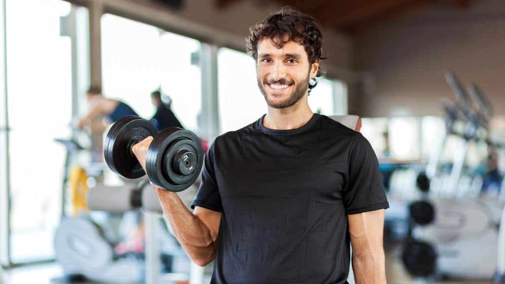 A man with curly hair smiles while performing a bicep curl with a black dumbbell.