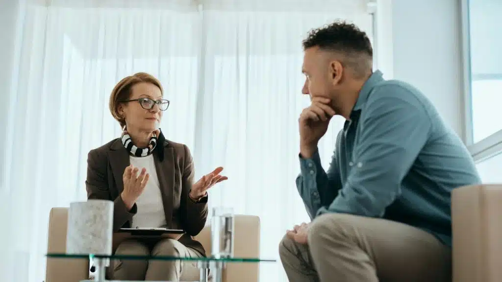 A woman with glasses speaks to a man while holding a clipboard in an office.