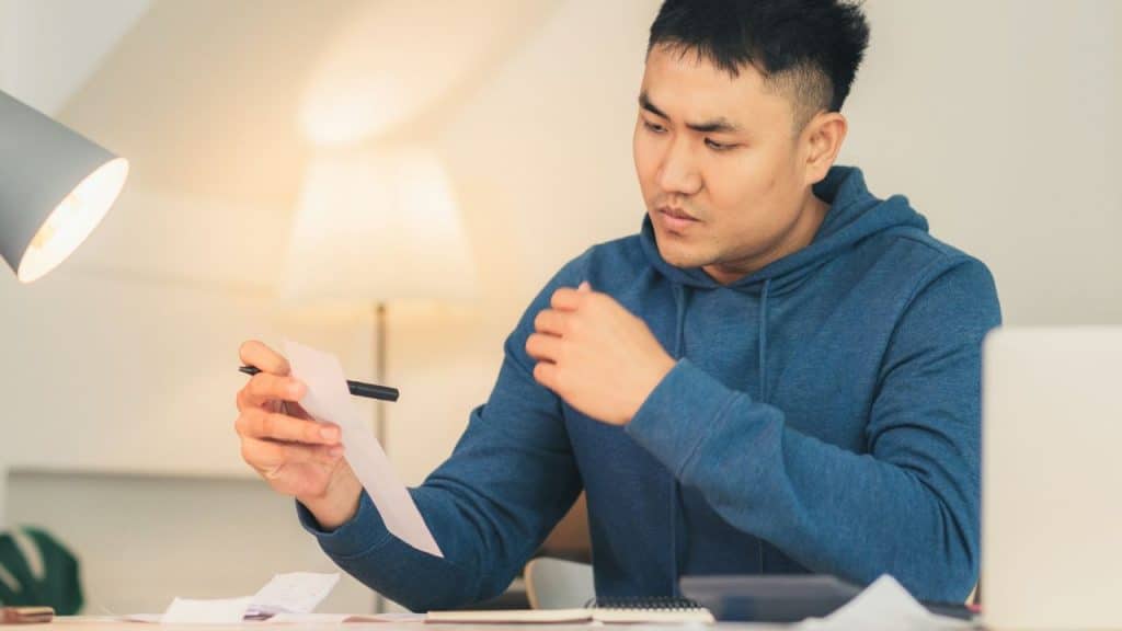 A man in a blue hoodie sits at a desk while looking at a white paper.