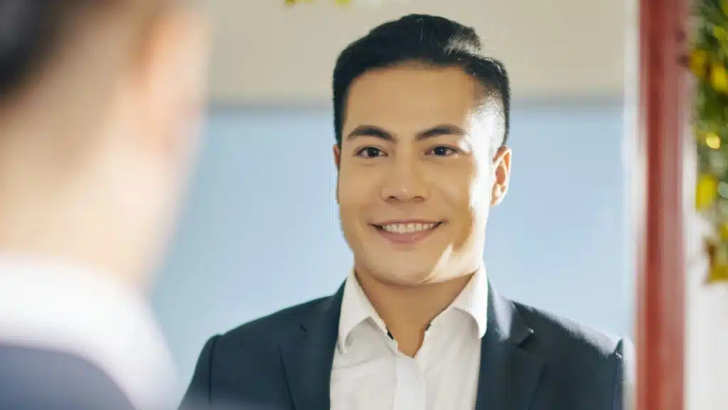 A smiling man in a suit and white shirt looks at his reflection in a mirror.