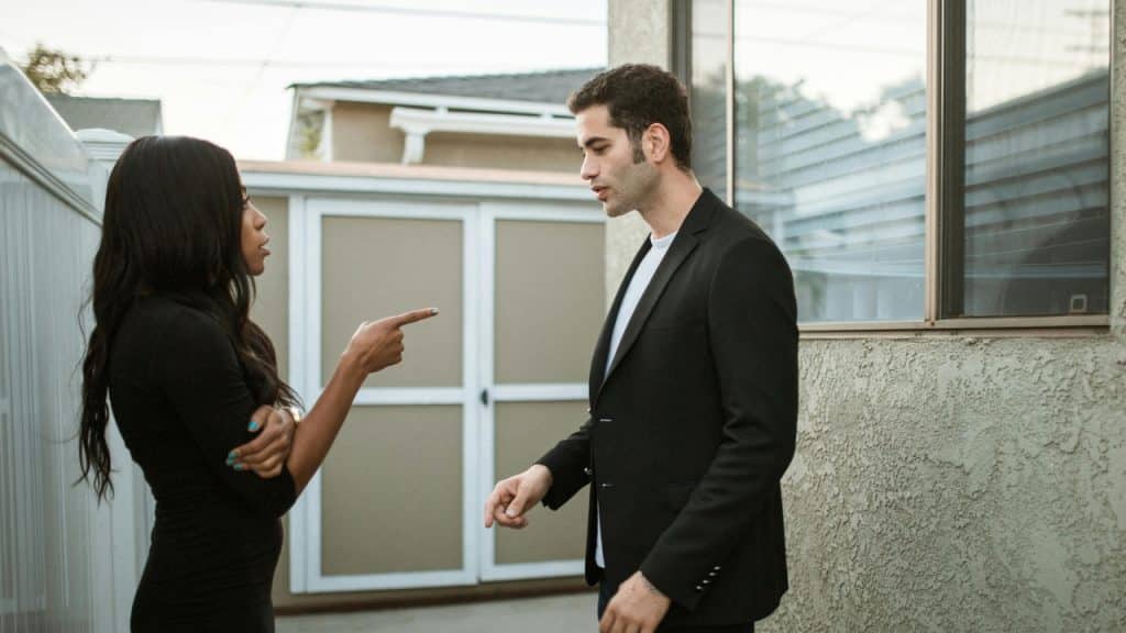 A woman points a finger toward a man standing outside near a light-colored wall.
