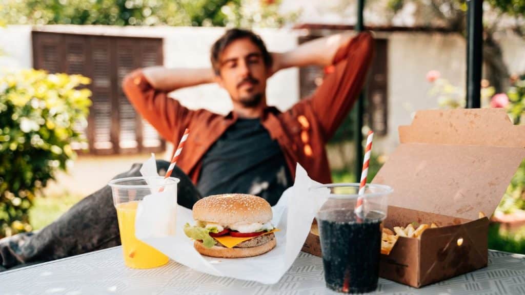 A man relaxes behind a table with a burger, fries, and two drinks.