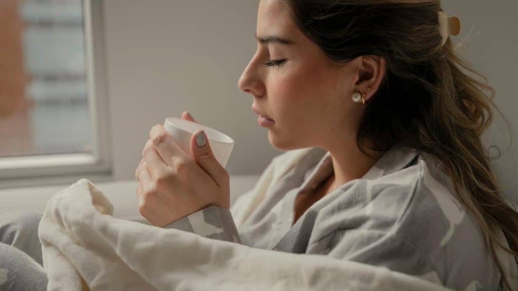 A woman wrapped in a blanket holding a warm drink by a window.