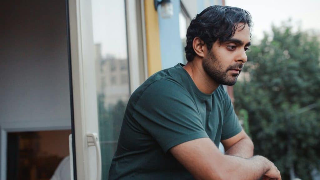 A man in a dark green shirt leans on a balcony railing and looks off into the distance.
