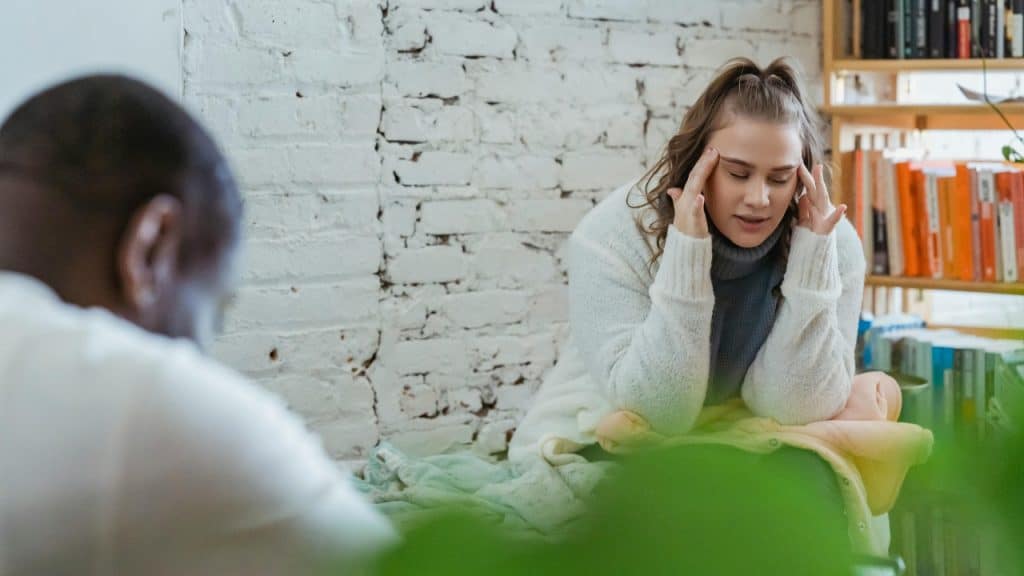 A woman sits with her eyes closed, holding her hands to her head in distress.