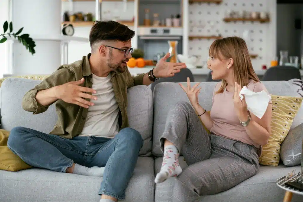 A man sits on a couch looking at a smartphone while holding a video game controller.
