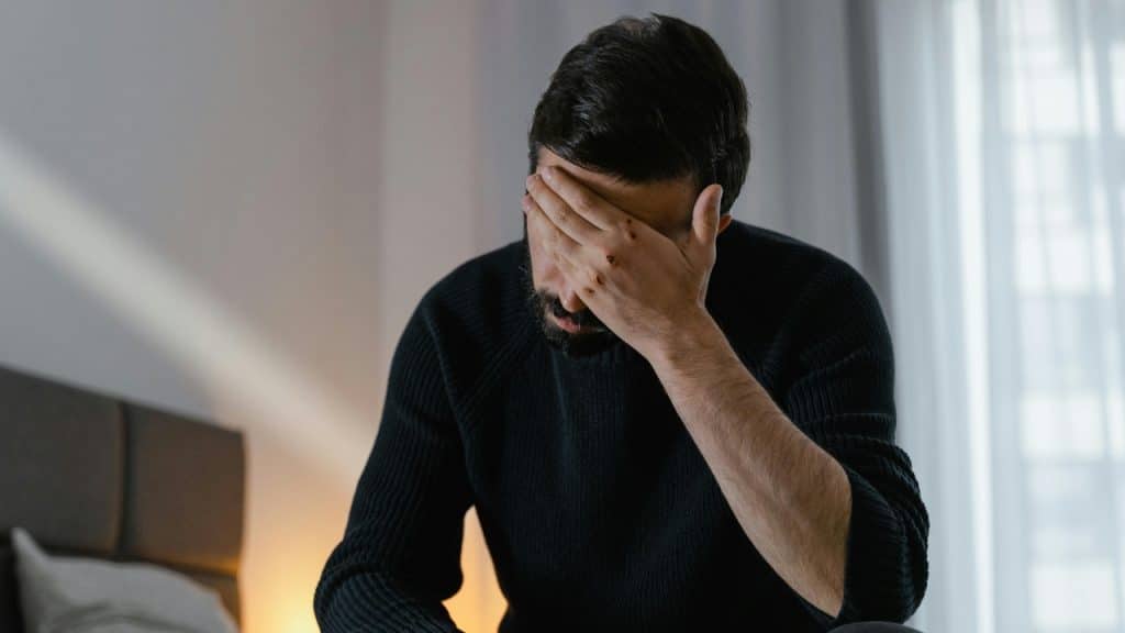 A man in a dark sweater sits on a bed, covering his eyes with his hand.