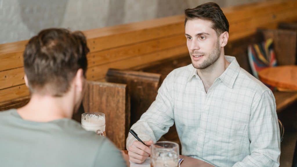 A man holding a pen sits across from another person at a table.