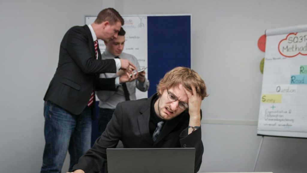 A man sits at a laptop looking frustrated while two colleagues talk in the background.