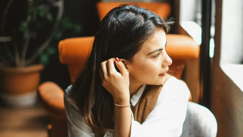 A woman sitting indoors in an orange chair, looking out a window while touching her hair.