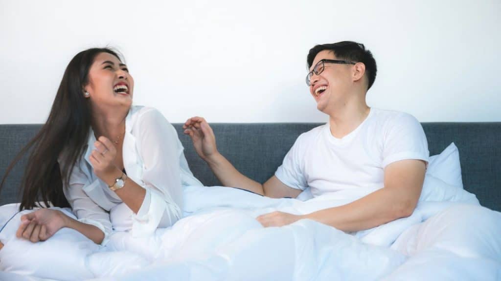 A laughing man and woman sit up in bed with white linens and a gray headboard.