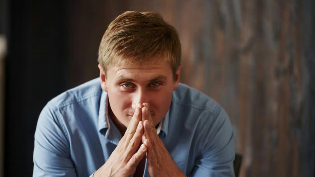 A man smiling at the camera and holding his hands upto his face with the fingers touching.