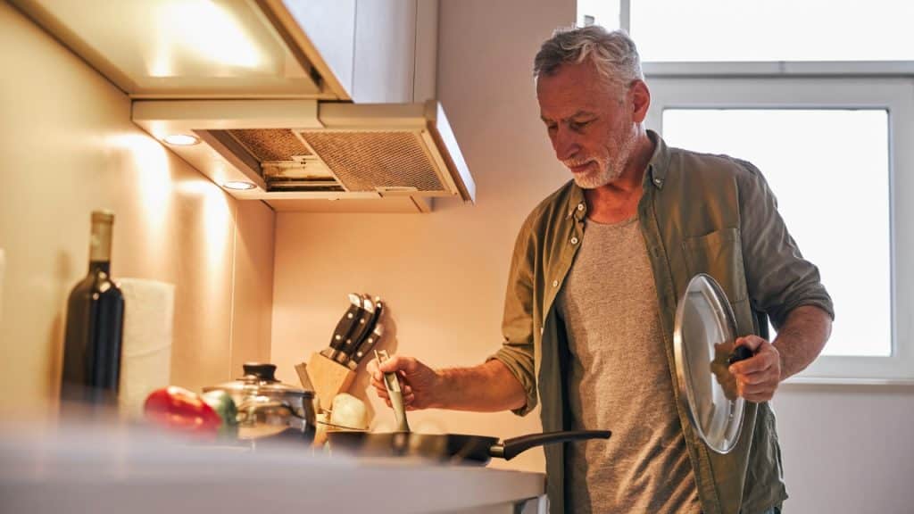 An older man in a green shirt stands in a kitchen stirring a frying pan.