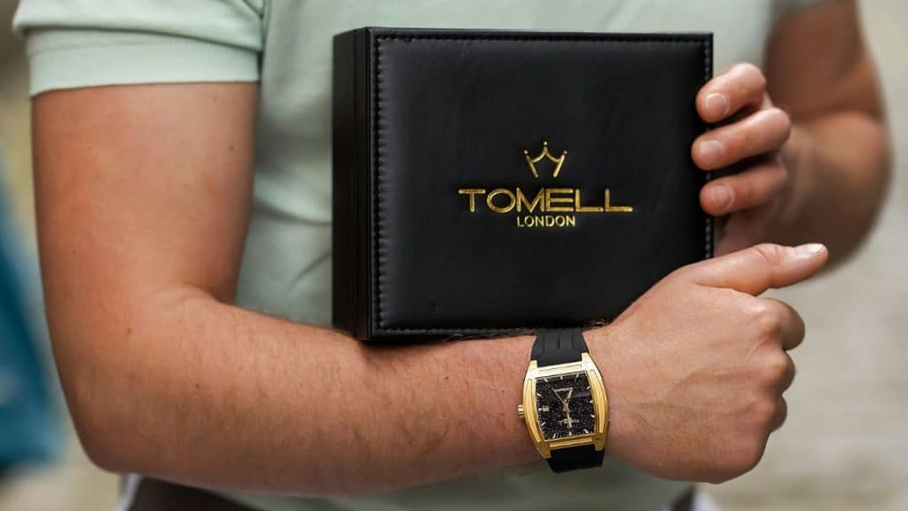 A person holds a Tomell London box while wearing a gold and black wristwatch.