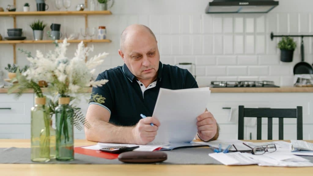 A man sits at a kitchen table reviewing paperwork with a calculator and glasses.