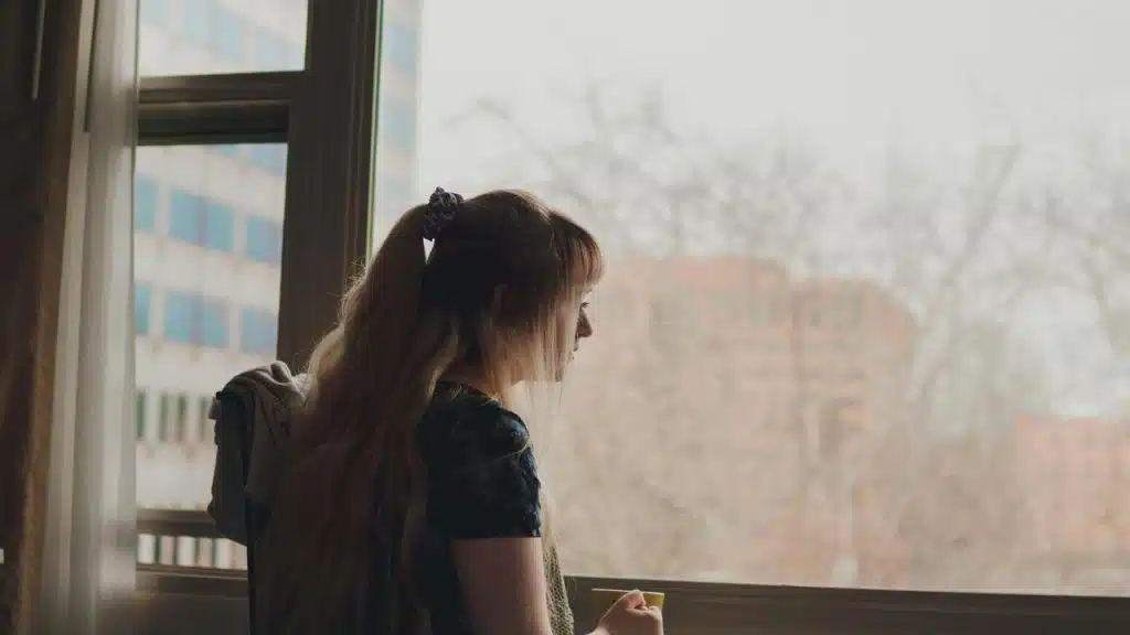 A woman standing by a window looking outside while holding a cup.